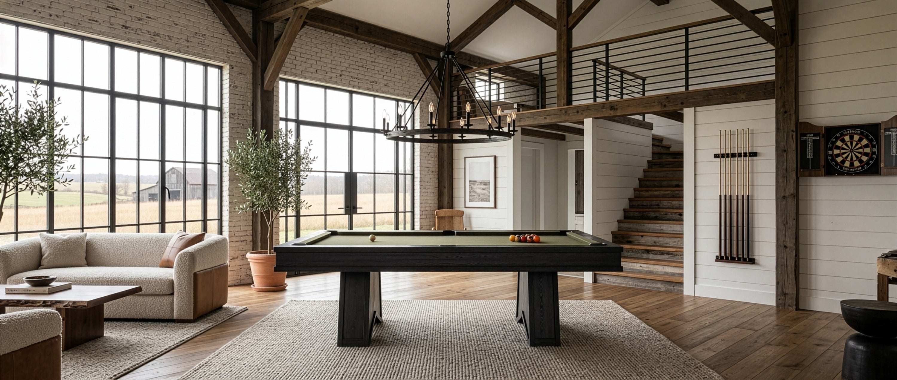 A modern dark wood pool table with a green felt surface is centered in a spacious, high-ceilinged rustic game room with exposed wood beams, large windows, a staircase, and a loft.