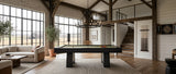 A modern dark wood pool table with a green felt surface is centered in a spacious, high-ceilinged rustic game room with exposed wood beams, large windows, a staircase, and a loft.