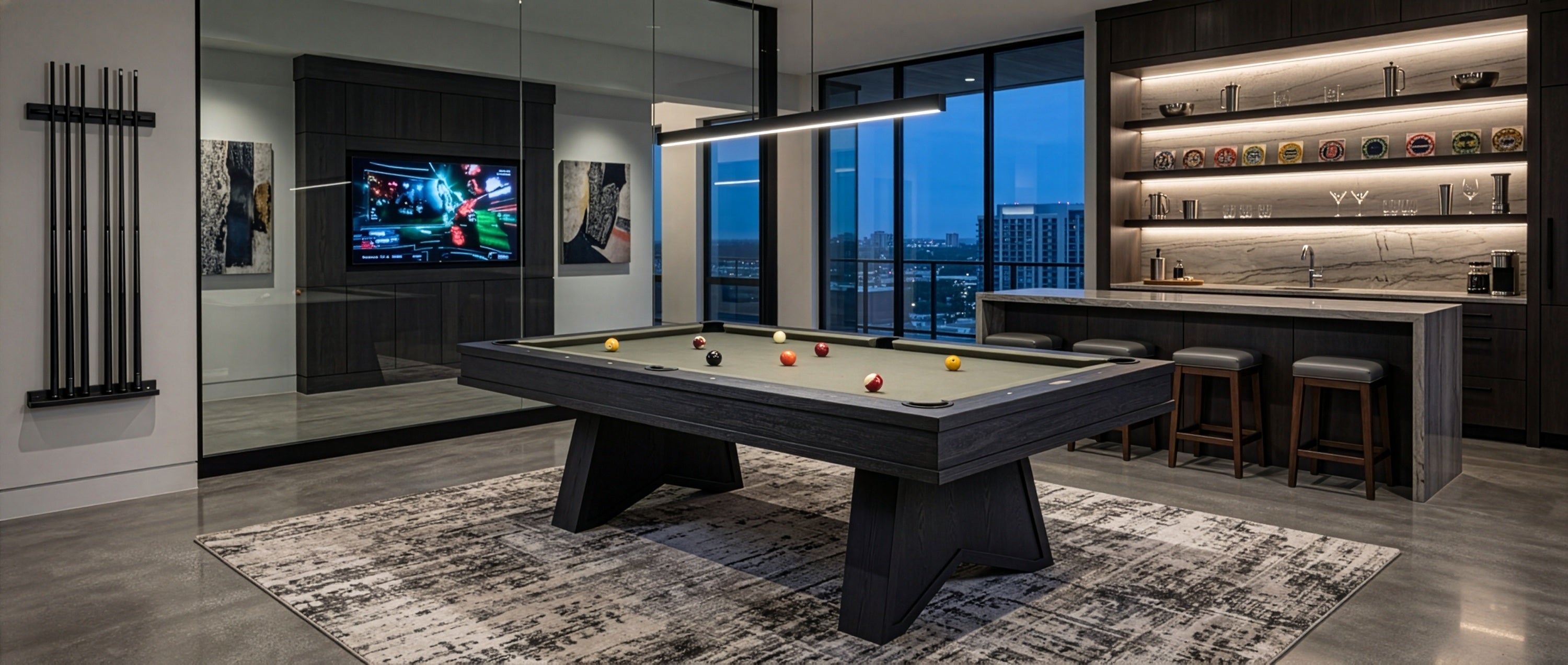 A modern pool table in a sleek, contemporary home bar or game room with dark wood cabinetry, built-in shelves, bar stools, a large wall-mounted TV, and floor-to-ceiling windows overlooking a city skyline at dusk.