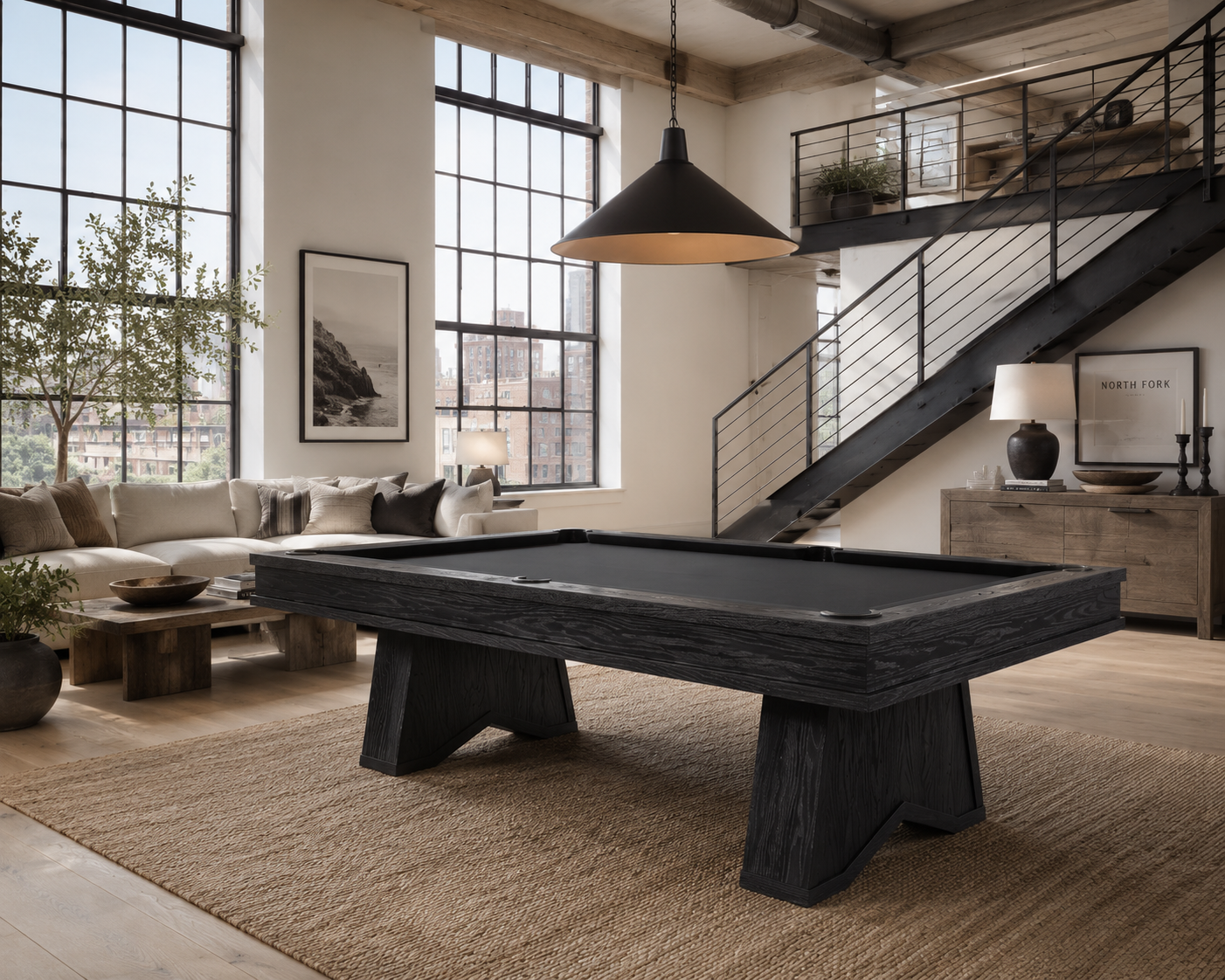 A dark wood-finish pool table with a black felt surface is shown in a modern loft-style living room with large windows, a staircase, and a beige rug.