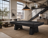 A dark wood-finish pool table with a black felt surface is shown in a modern loft-style living room with large windows, a staircase, and a beige rug.