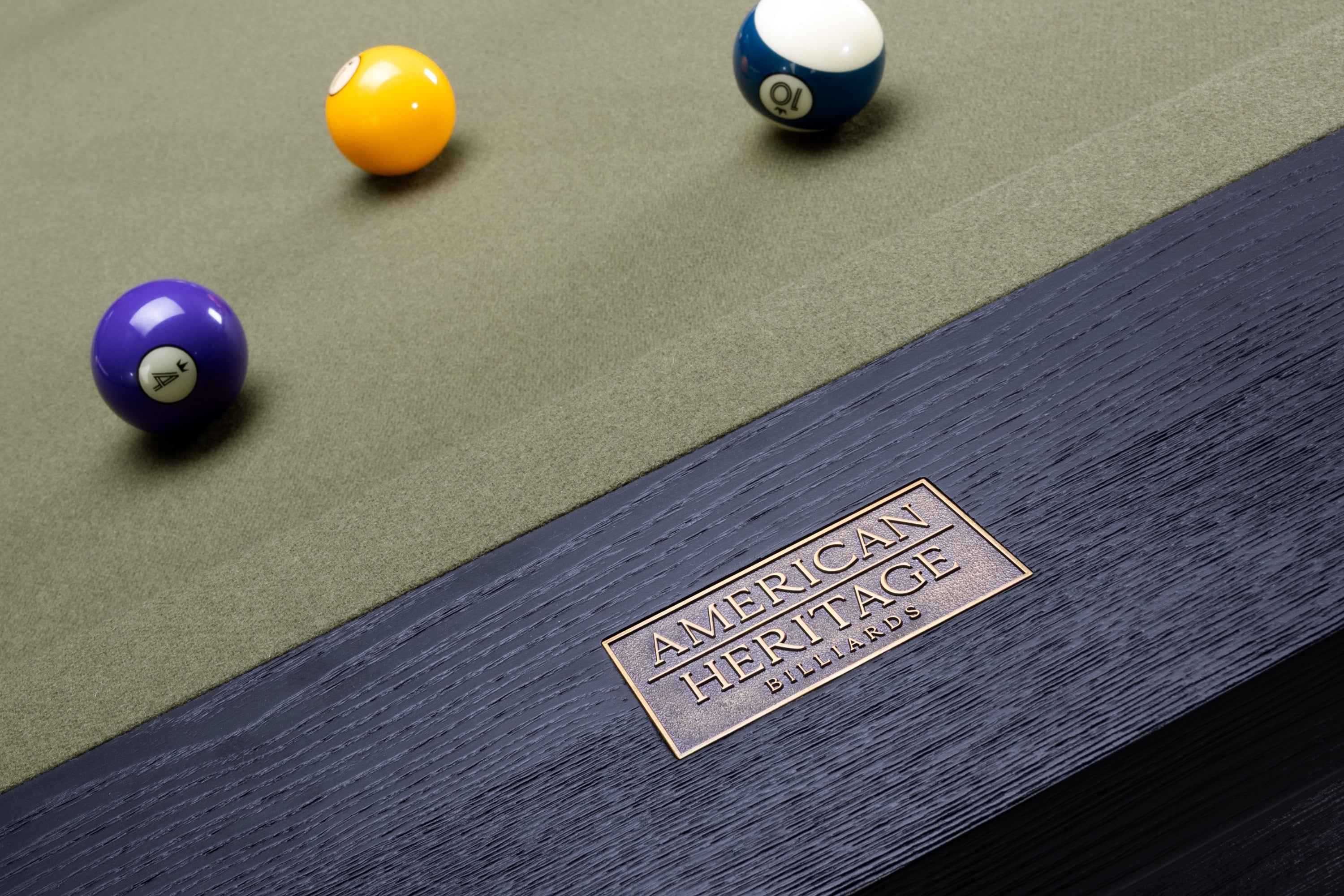 Close-up of a wooden billiards table corner with a brass plaque reading American Heritage Billiards, a green felt surface, and three pool balls nearby.