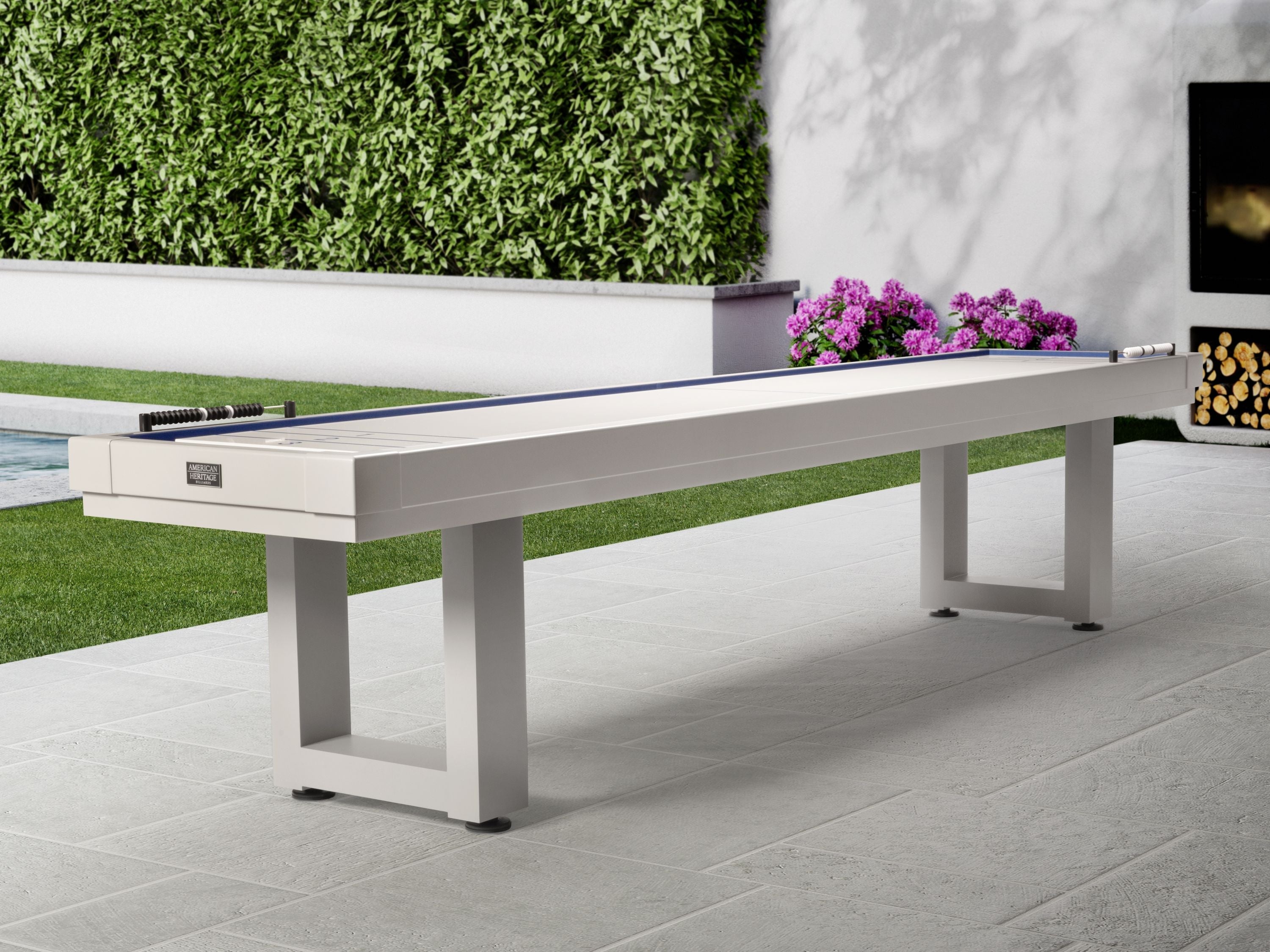An American Heritage Lanai 9' Pearl White shuffleboard table stands by a hedge and flowers.