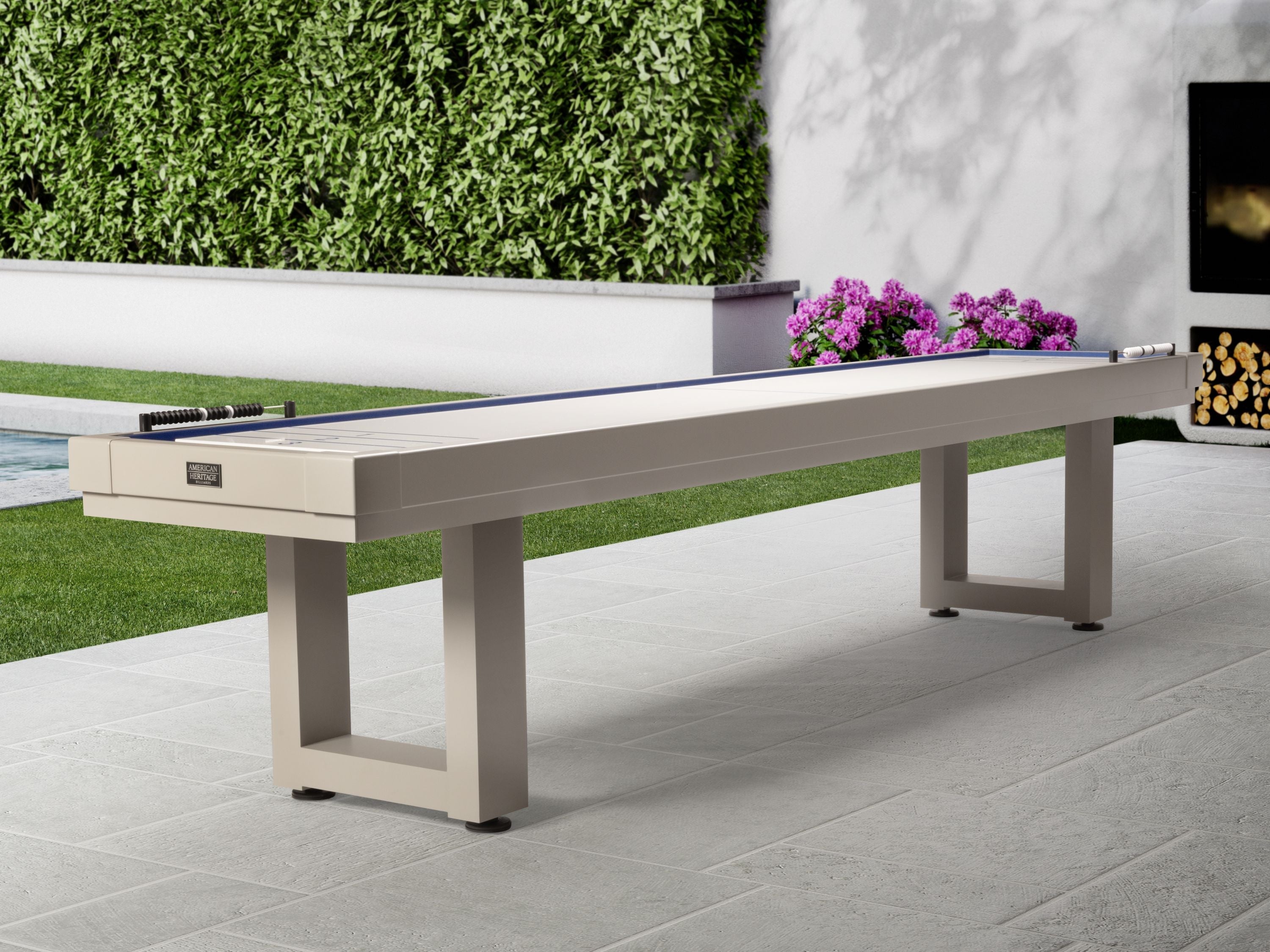 American Heritage Lanai 12' Outdoor Shuffleboard Table in Oyster Grey on a stone patio with flowers.