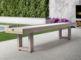 American Heritage Lanai 12' Outdoor Shuffleboard Table in Oyster Grey on a stone patio with flowers.