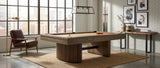 A modern wooden pool table in a bright, contemporary loft room with large industrial windows, a brown leather armchair, and a white brick wall.