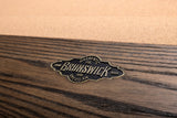 Close-up of a Brunswick hardwood pool table finish showing a dark brown wood grain surface with a brass logo plaque reading 'Authentic Brunswick 1965 American'.